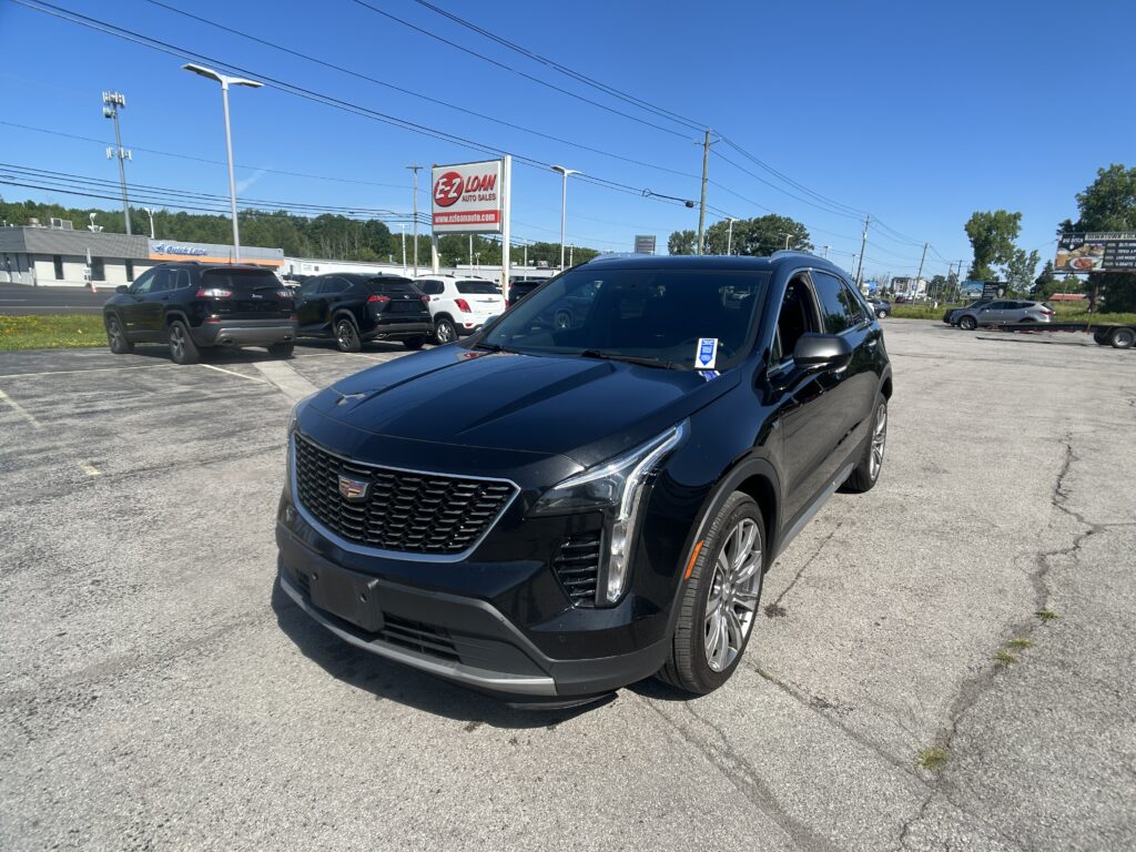 Black Cadillac SUV in the foreground at a car dealership lot with an EZ Loan Auto Sales sign and other vehicles in the background under a clear blue sky.