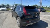 Rear view of a shiny black Cadillac XT4 SUV in a car dealership lot with other vehicles and a clear blue sky above.