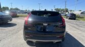 Rear view of a black Cadillac XT4 SUV in a car dealership lot, surrounded by other parked cars under a clear blue sky, with power lines overhead.