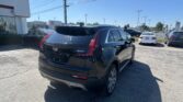 Rear three-quarter view of a black Cadillac SUV in a car dealership lot on a sunny day, with many cars and Toyota signs in the background.