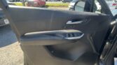 Interior view of a car door panel with a chrome door handle and window/lock controls; outside is a busy dealership parking lot visible through the window