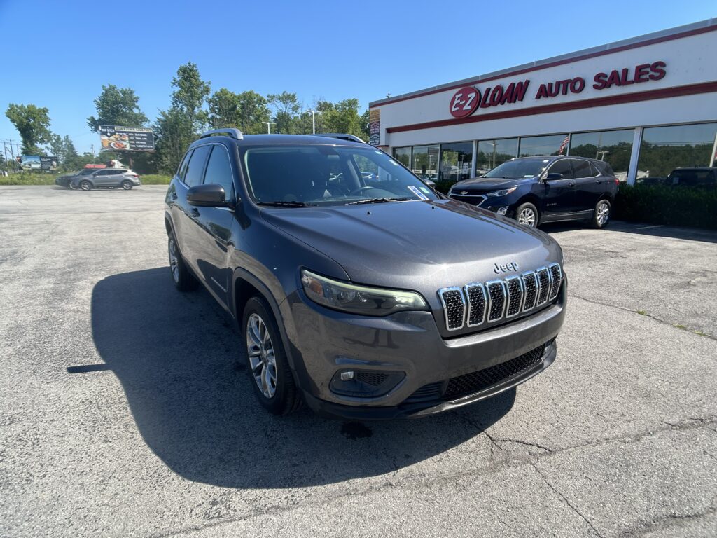 Grey Jeep SUV in foreground at a car lot with 'EZ Loan Auto Sales' building in background and clear blue sky