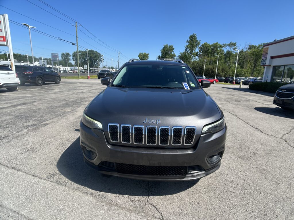 Front view of a dark gray Jeep SUV parked in a sunny car dealership lot with other vehicles nearby.