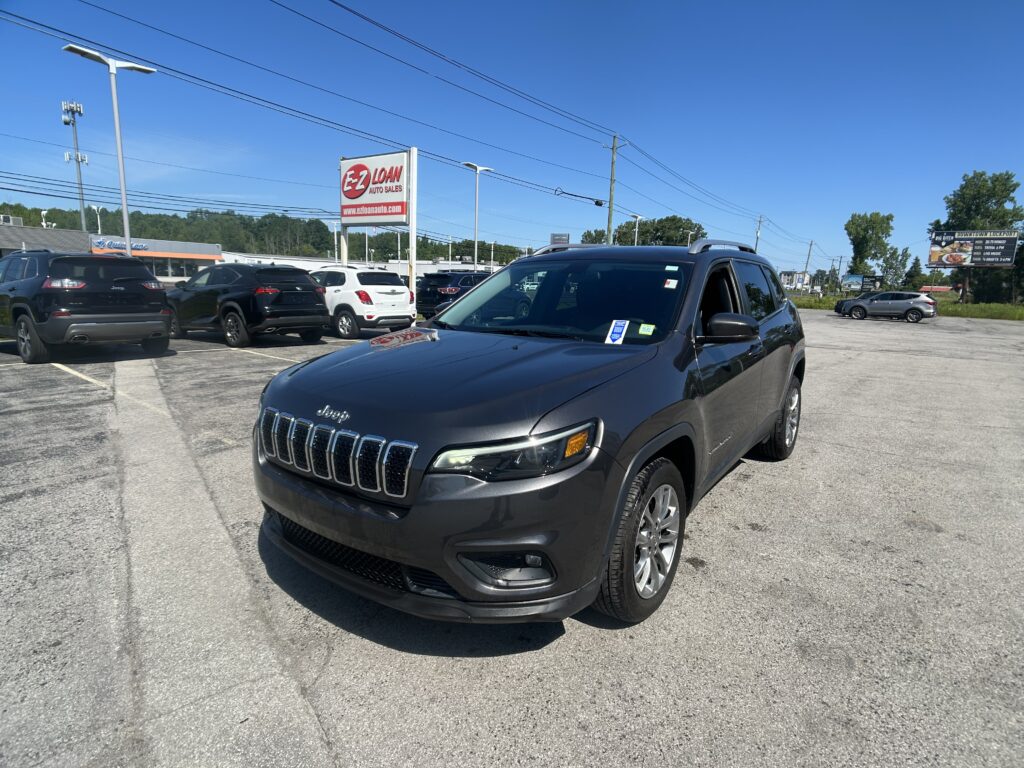 Close-up of a dark gray Jeep SUV in a car lot with other vehicles and an EZ LOAN Auto Sales sign in the background under a clear blue sky.