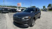 Close-up of a dark gray Jeep SUV in a car lot with other vehicles and an EZ LOAN Auto Sales sign in the background under a clear blue sky.