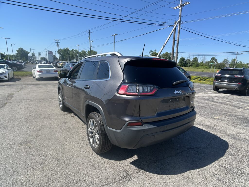 Rear three-quarter view of a gray Jeep SUV in a busy outdoor parking lot on a sunny day with power lines overhead.