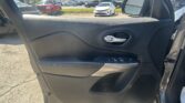 Interior of a car door showing the black textured panel, chrome door handle, and window controls, with a parking lot visible outside the open doorway and side mirror reflecting cars.