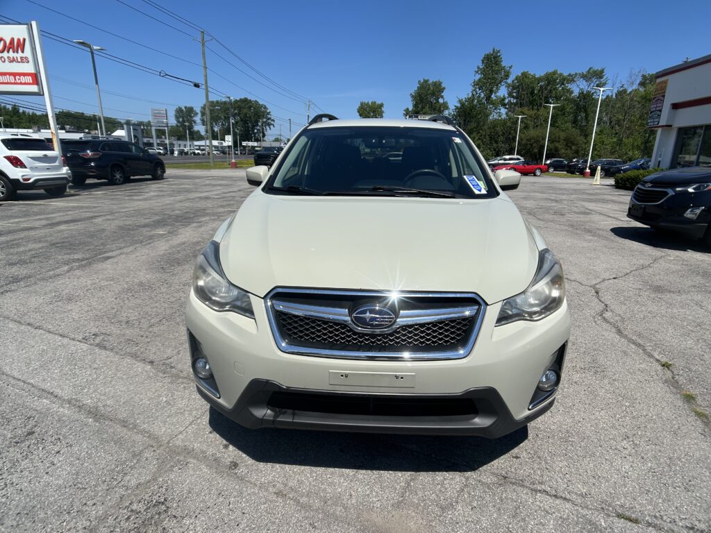 Front view of a white Subaru SUV parked in a car dealership lot on a sunny day, with other vehicles and a sign visible in the background.