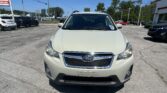 Front view of a white Subaru SUV parked in a car dealership lot on a sunny day, with other vehicles and a sign visible in the background.