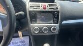 Car interior center console: touchscreen radio display, climate dials, and hazard switch, seen from driver's seat with outside parking lot glare through windshield.