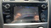 Center console in a car: touchscreen showing rear‑view camera with yellow/green/red guide lines and message 'Check Surroundings Before Backing Up'; hazard light button above screen; volume and tuning dials on either side.