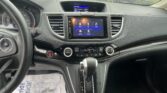 Driver's-eye view of a car interior: steering wheel left, central touchscreen infotainment system, and gear shifter visible atop a dark dashboard with a rain-speckled windshield outside.