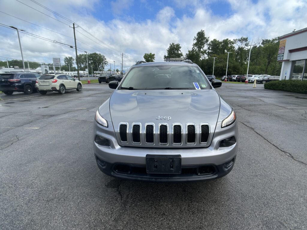 Front view of a silver Jeep Grand Cherokee parked in a dealership lot with other cars and a building in the background under a partly cloudy sky.