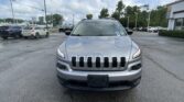 Front view of a silver Jeep Grand Cherokee parked in a dealership lot with other cars and a building in the background under a partly cloudy sky.
