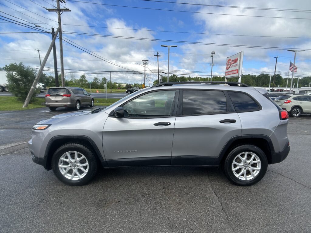 Silver Jeep Grand Cherokee in a car lot with overhead power lines, a dealership sign reading 'EZ Loan Auto Sales' in the background, and an American flag nearby.