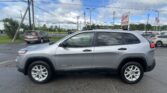 Silver Jeep Grand Cherokee in a car lot with overhead power lines, a dealership sign reading 'EZ Loan Auto Sales' in the background, and an American flag nearby.