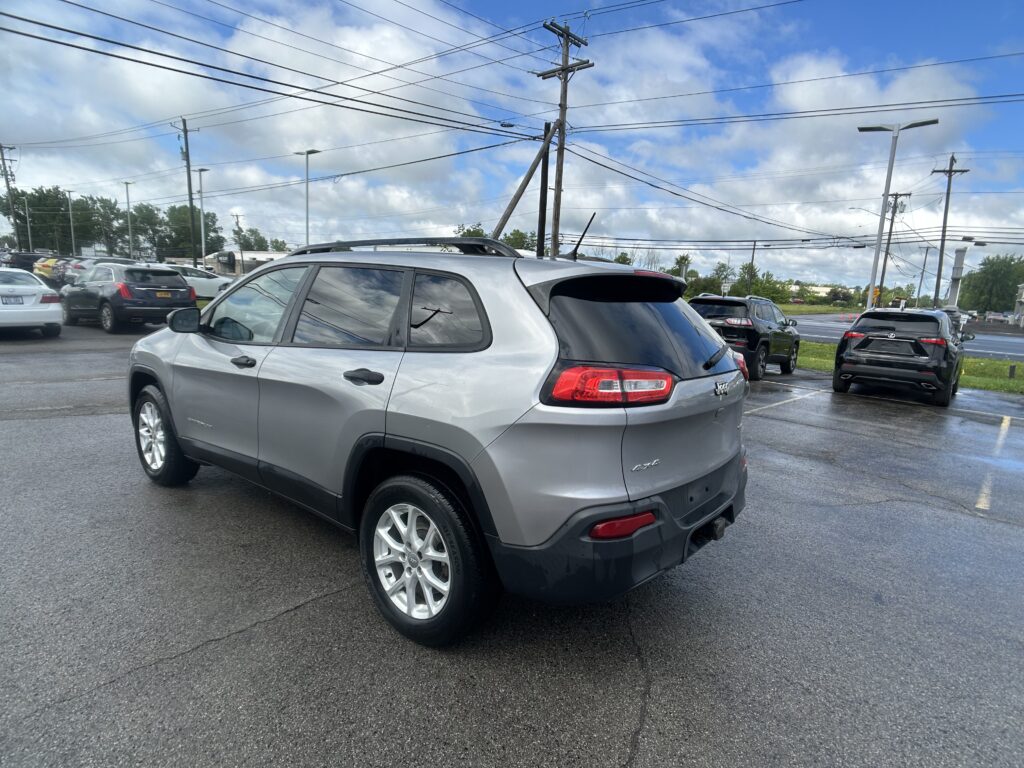 Silver Jeep SUV parked in a wet lot with other cars, power lines overhead and a blue, cloudy sky above.