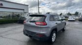 Rear view of a silver Jeep SUV in a car dealership lot, with a white building on the left and rows of cars under a cloudy sky.