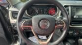 Driver's view of a Jeep dashboard with a circular speedometer and tachometer, steering wheel, and center console; outside is a parking lot seen through the windshield.