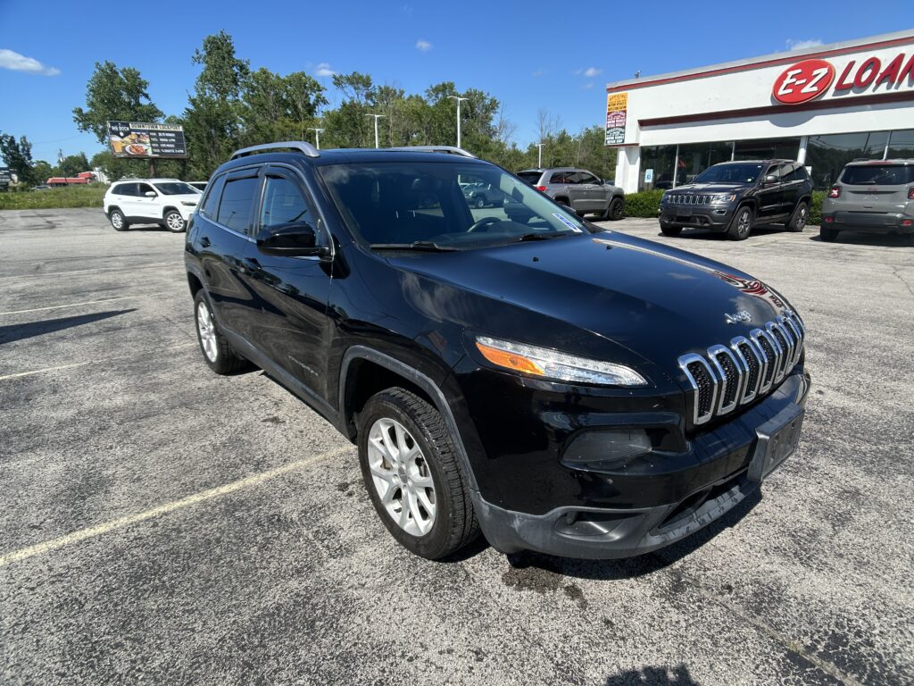 Black Jeep Grand Cherokee parked diagonally in a dealership lot with other cars and a building in the background on a sunny day.