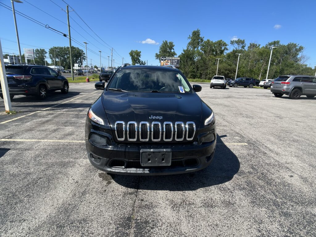 Front view of a dark Jeep SUV in a parking lot with other cars and a clear blue sky behind it.