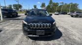 Front view of a dark Jeep SUV in a parking lot with other cars and a clear blue sky behind it.