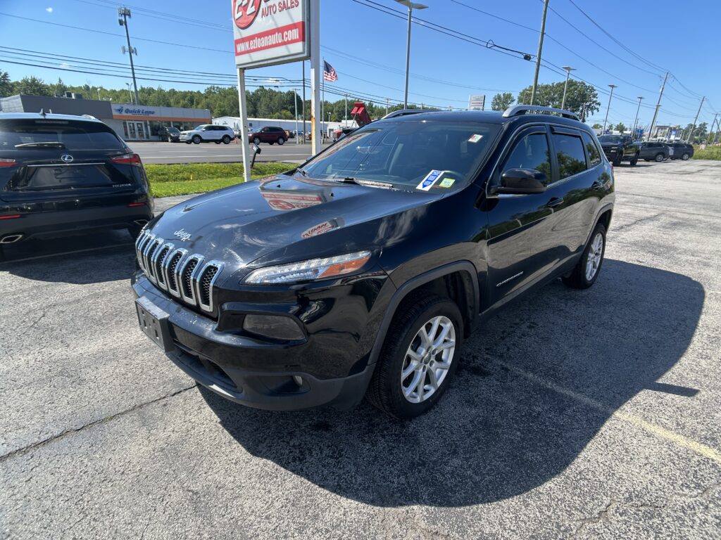 Black Jeep Grand Cherokee in the foreground at a car dealership lot with other vehicles and a tall sign in the background under a blue sky.
