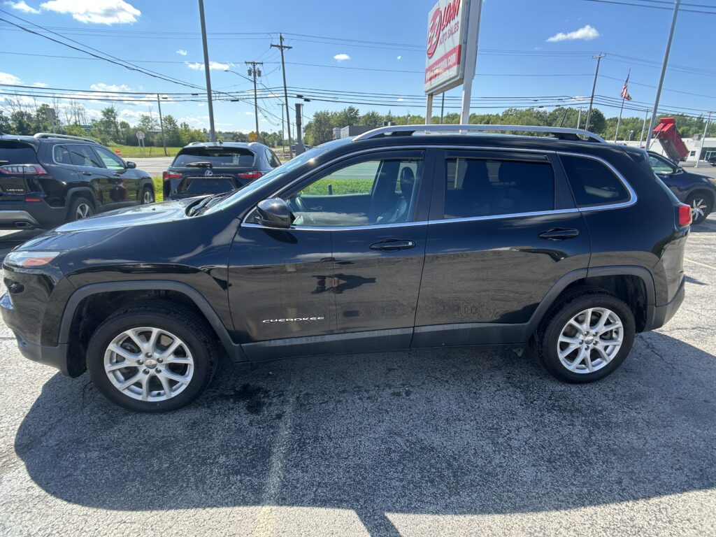 Black Jeep Cherokee SUV parked in a car lot with other vehicles and a tall sign in the background on a sunny day.