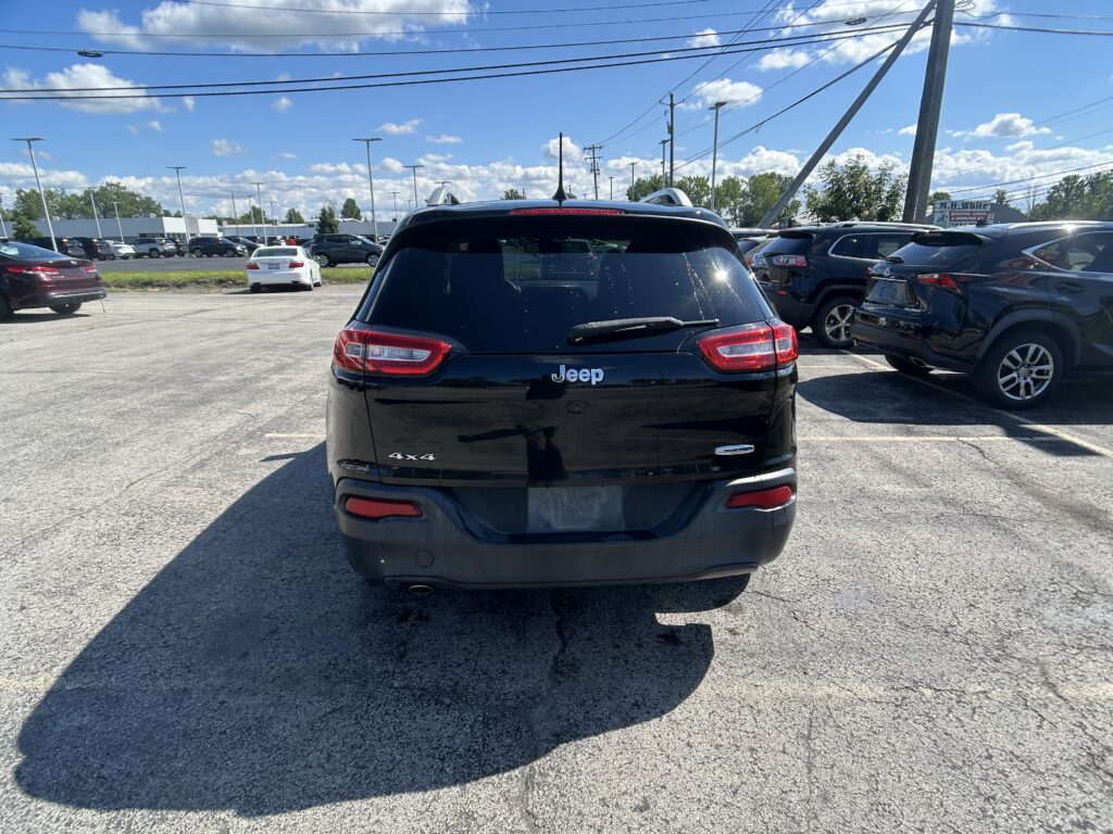 Rear view of a black Jeep SUV in a crowded dealership lot with other cars and power lines overhead under a blue sky with clouds