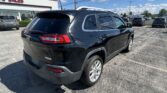 Black Jeep SUV parked in a car dealership lot on a sunny day, with other cars and a white Auto Sales building in the background.