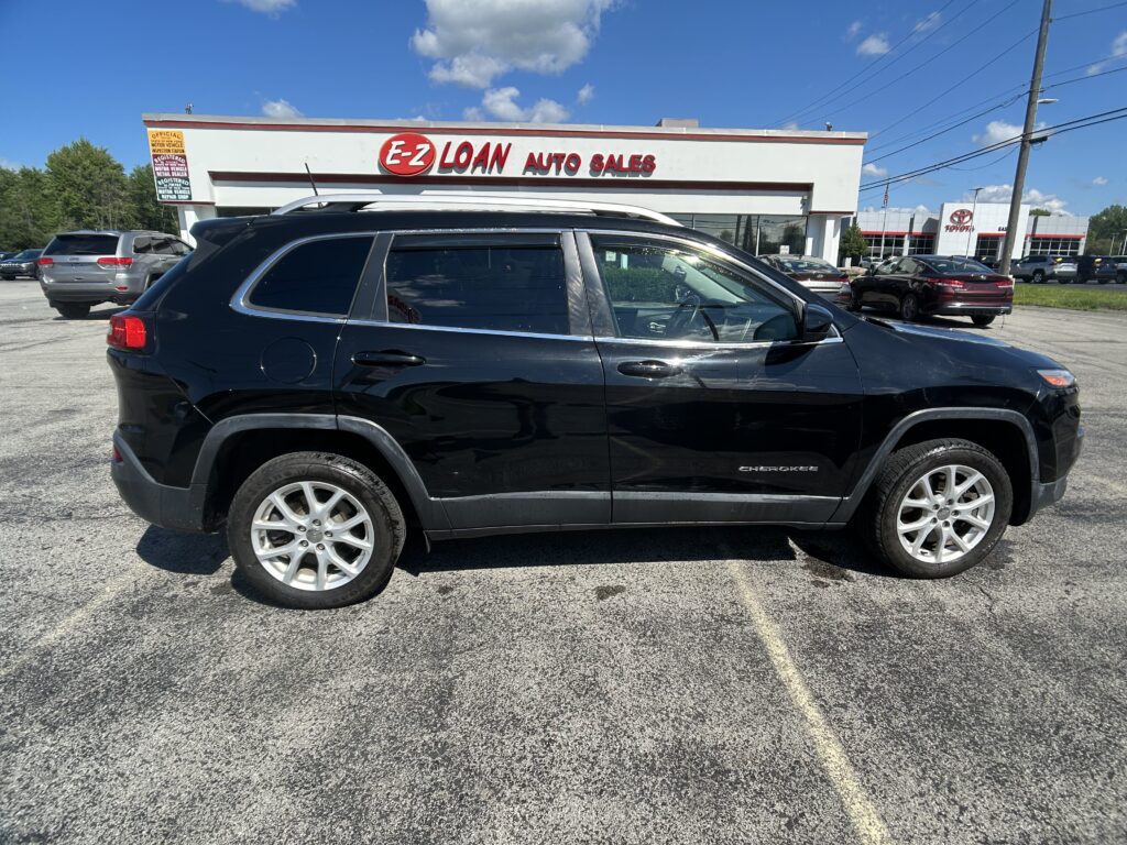 Black Jeep Cherokee SUV in the foreground with EZ Loan Auto Sales storefront and a bright blue sky in the background.