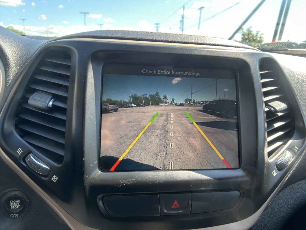 Car dashboard touch screen showing rearview camera with parkingGuidance lines; message reads 'Check Entire Surroundings' and the view of a parking lot ahead