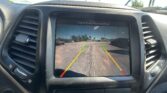 Car dashboard touch screen showing rearview camera with parkingGuidance lines; message reads 'Check Entire Surroundings' and the view of a parking lot ahead