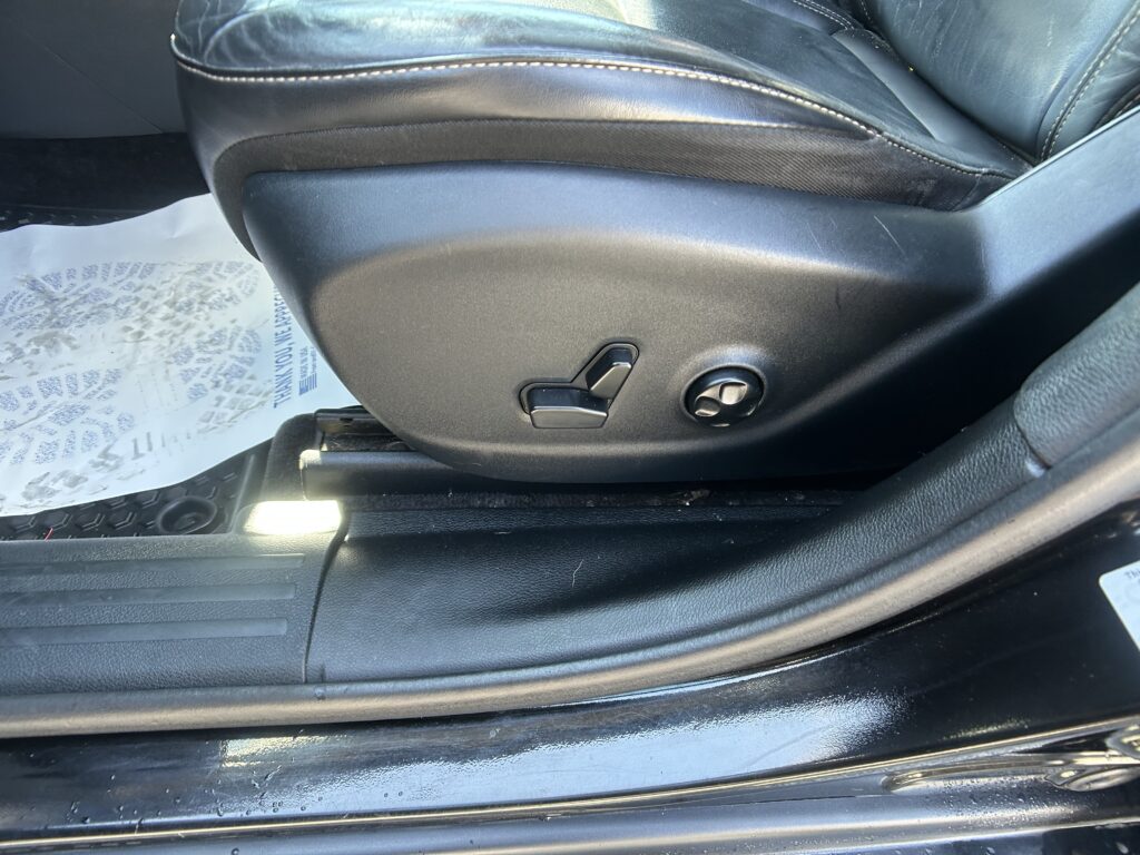 Driver-side car seat with black leather upholstery and power-adjustment controls, including lumbar support lever and seat height knob, on a clean interior panel in daylight.