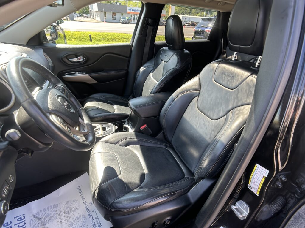 Dark leather car interior with Jeep branding on the front seats, shown from the driver's side, including steering wheel, center console, and outside street view through the windows.