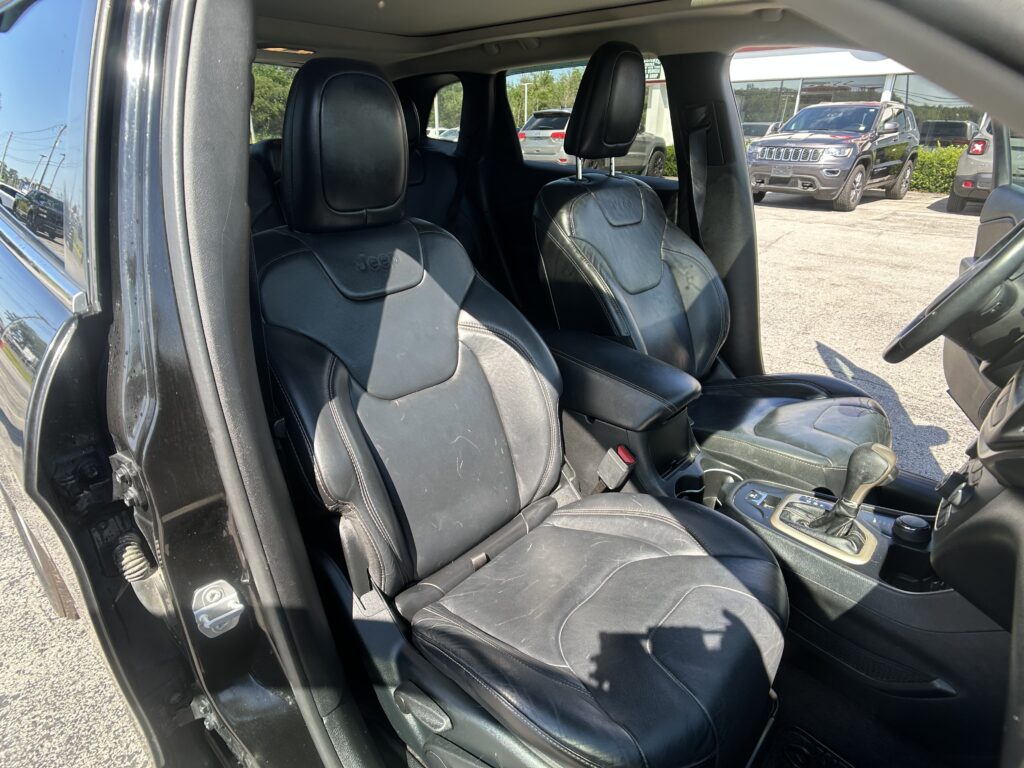 Front row of a black leather SUV interior with driver and passenger seats, center console visible, dealership lot outside the open door.