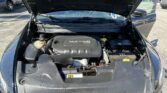 Under-hood view of a dark SUV with the hood raised, exposing the engine and battery in a parking lot