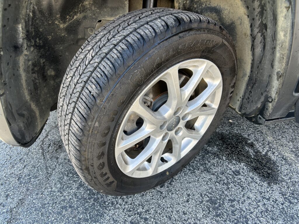 Close-up of a car tire and alloy wheel with dirt-splattered wheel well, resting on a paved surface.