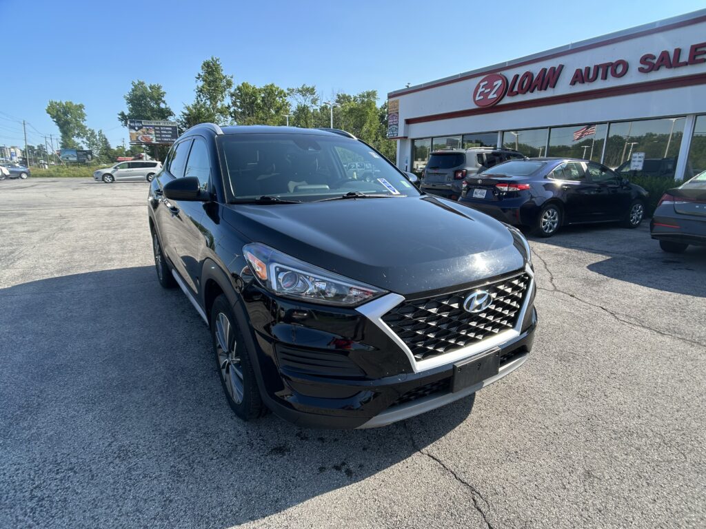 Front corner of a black Hyundai SUV parked in a dealership lot with a white building and 'E-Z LOAN AUTO SALES' sign in the background.