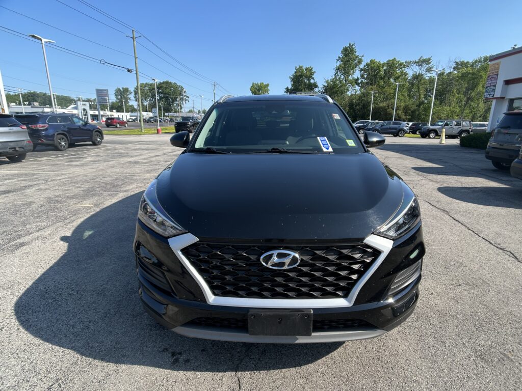 Close-up of a black Hyundai SUV front in a parking lot, with other cars and a dealership building in the background under a clear blue sky.