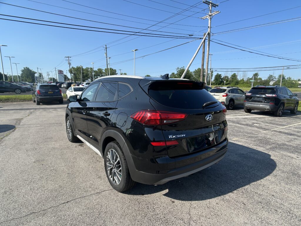 Rear three-quarter view of a black Hyundai Tucson parked in a busy dealership lot with other cars and overhead power lines under a clear blue sky.