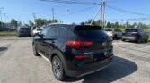 Rear three-quarter view of a black Hyundai Tucson parked in a busy dealership lot with other cars and overhead power lines under a clear blue sky.