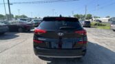 Rear view of a black Hyundai Tucson in a car dealership lot with other cars and overhead power lines under a clear blue sky.