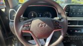 Driver’s-seat view of a car cockpit with a textured steering wheel, digital instrument cluster, and a touchscreen infotainment system showing radio; parking lot visible outside the windshield