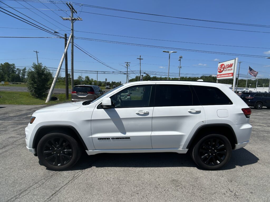 White SUV (Jeep Grand Cherokee) in a parking lot with utility poles and multiple power lines overhead; dealership sign and American flag visible in the background