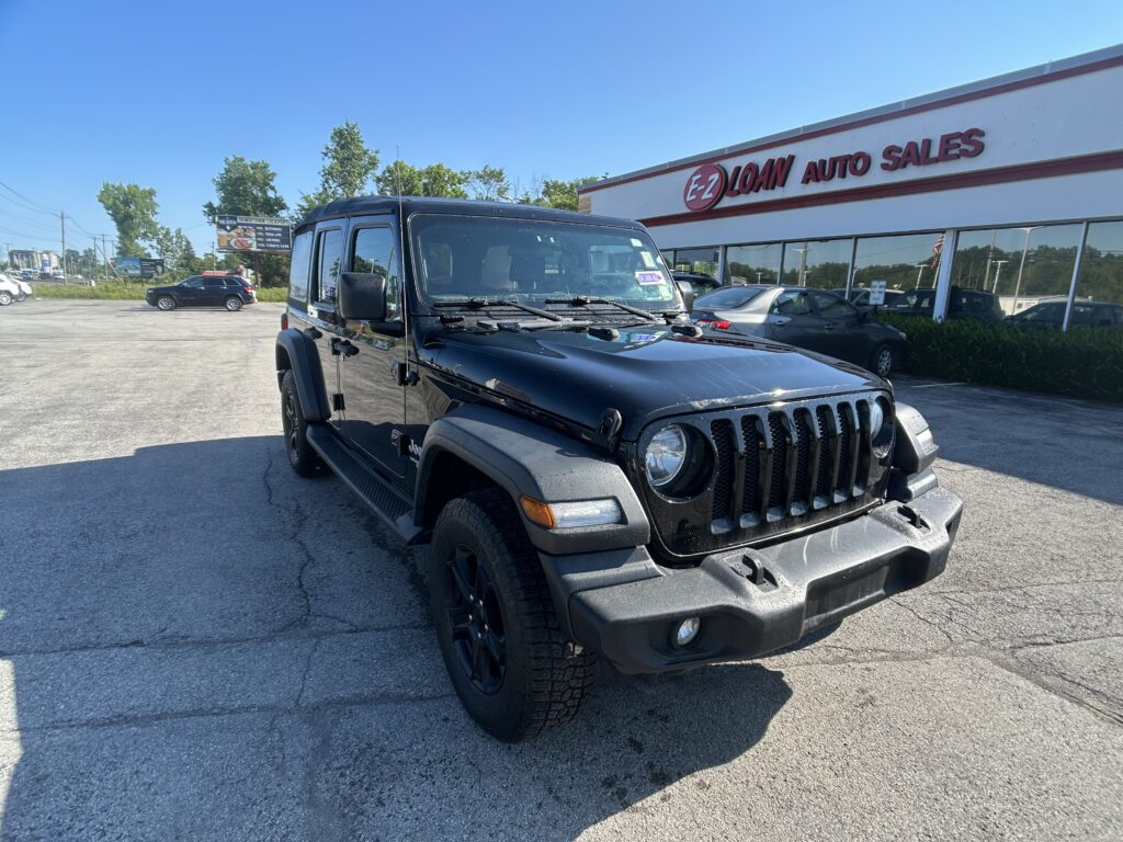 Black Jeep Wrangler in a dealership lot with EZ Loan Auto Sales sign on the building under a clear blue sky.