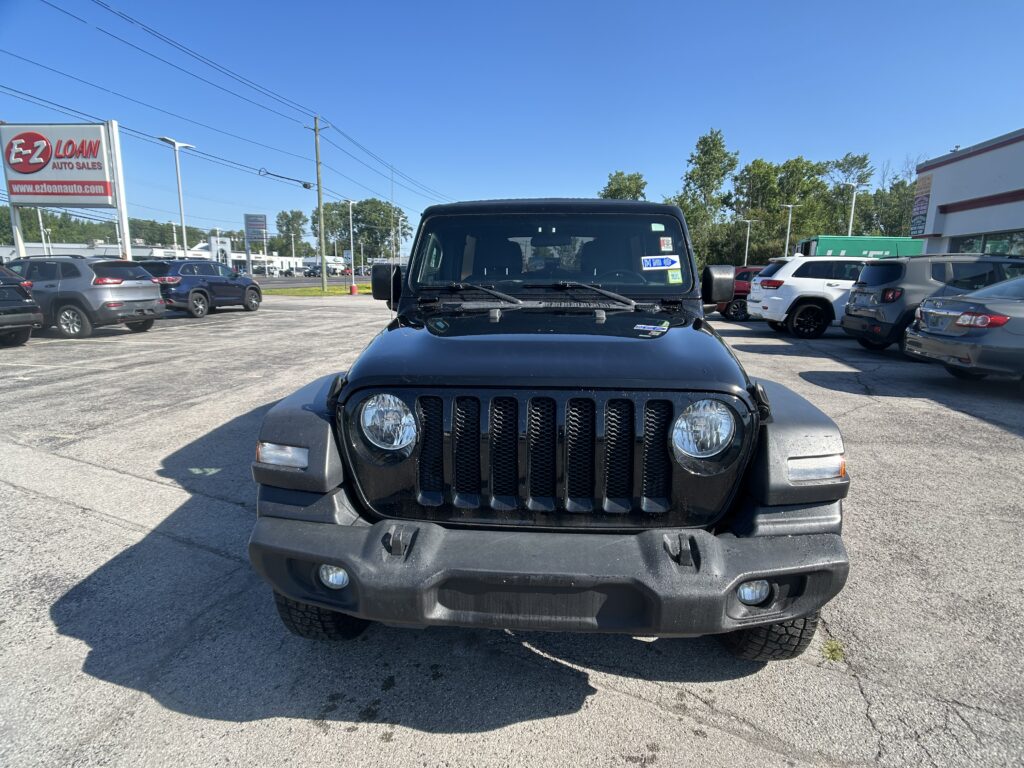 Front view of a black Jeep Wrangler parked in a dealership lot, with an EZ Loan Auto Sales sign visible on the left and other cars surrounding the lot under a clear blue sky.