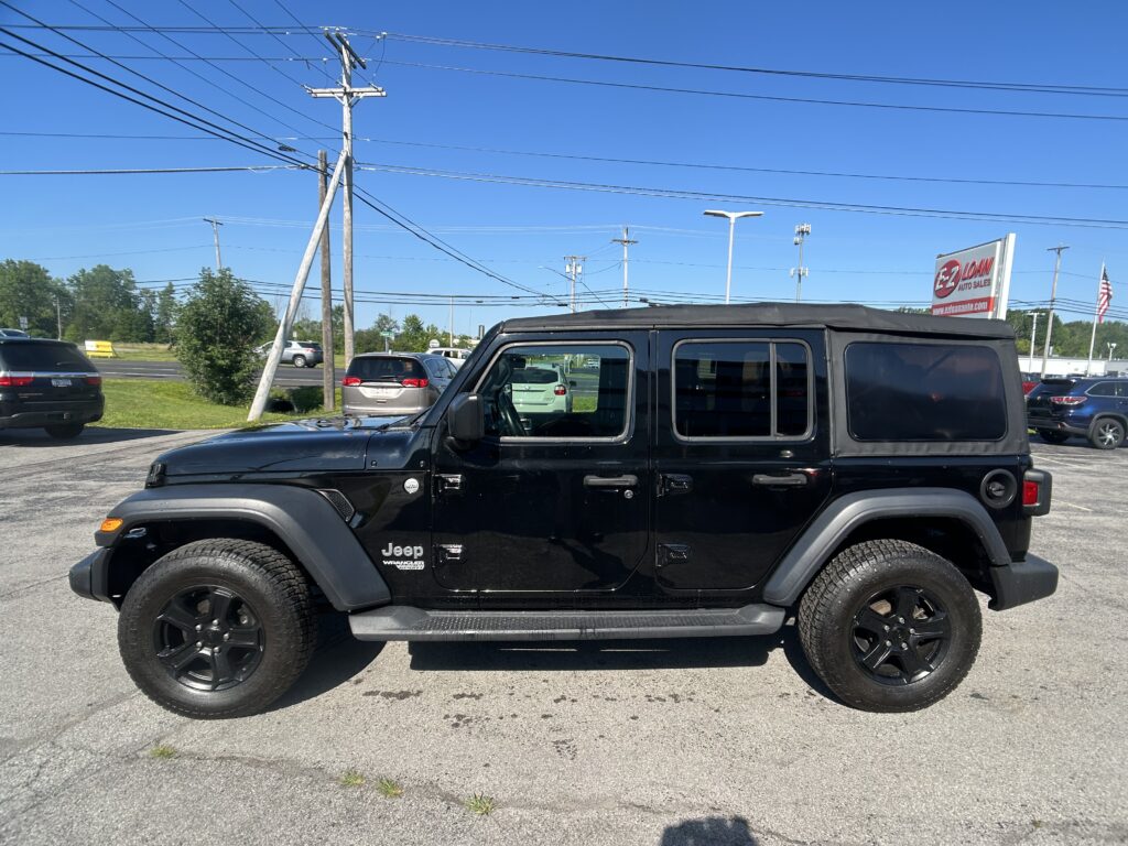 Black Jeep Wrangler parked in a dealership lot with power lines and a bright blue sky in the background, EZ Loan Auto Sales sign visible in the distance.
