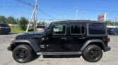 Black Jeep Wrangler parked in a dealership lot with power lines and a bright blue sky in the background, EZ Loan Auto Sales sign visible in the distance.