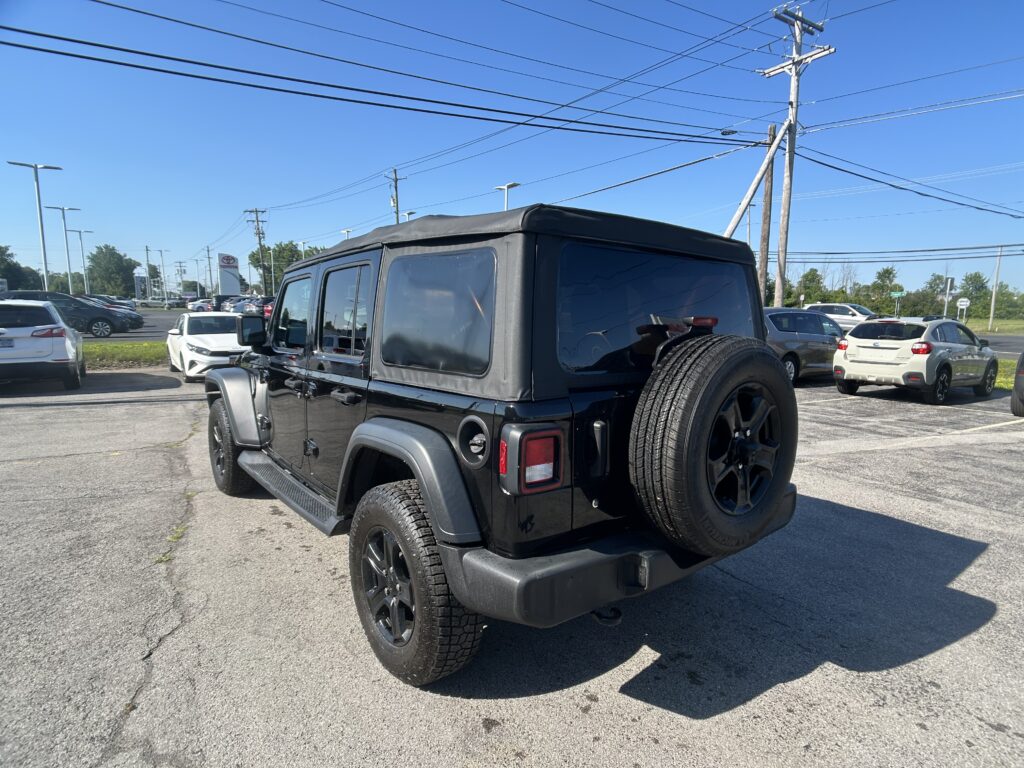 Rear three-quarter view of a black Jeep Wrangler with a soft top in a parking lot, spare tire on the back, blue sky above.